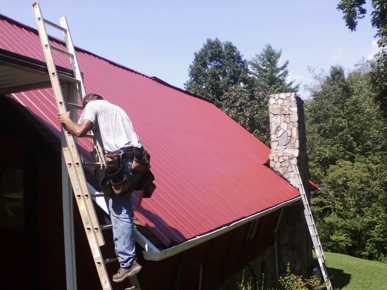 Roofer on a ladder beside a red metal roof and stone chimney — Soffit & Fascia Repair by Roofing Brothers in Carmichaels